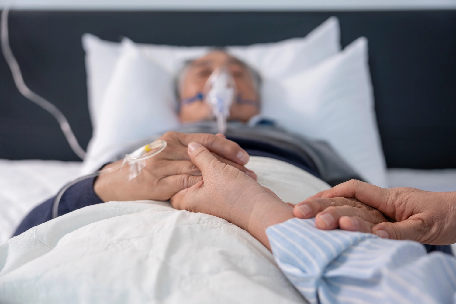 Curanus GmbH – Pflege – Szene: Familie hält die Hände eines sterbenden älteren Patienten, der mit Sauerstoff und Infusion im Bett liegt.