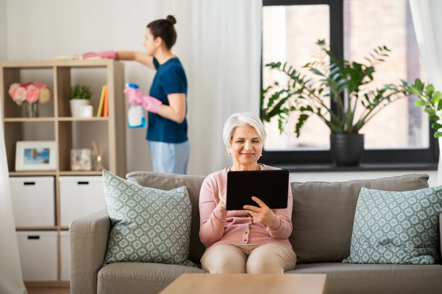 Curanus GmbH – Pflege – Technologie, Alter und Menschen – Glückliche ältere Frau mit Tablet-PC und Haushaltshilfe beim Putzen